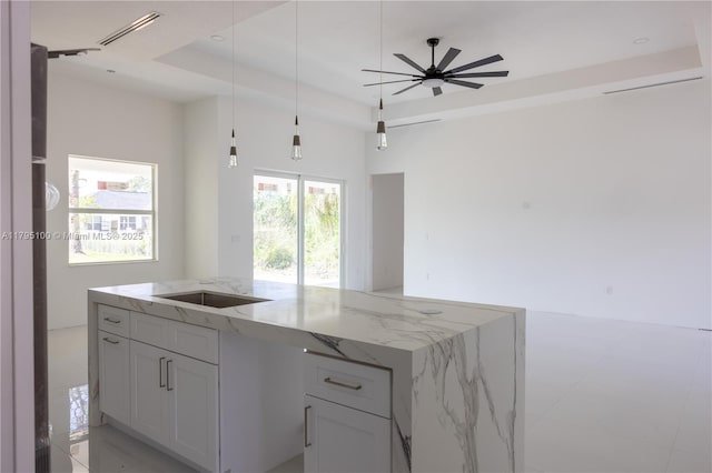 kitchen with hanging light fixtures, light stone counters, a raised ceiling, a kitchen island with sink, and open floor plan