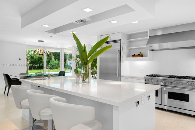 kitchen featuring wall chimney range hood, white cabinets, light stone counters, an island with sink, and premium appliances