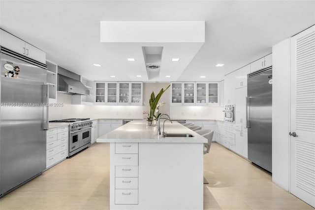 kitchen with high quality appliances, a center island with sink, white cabinetry, a sink, and wall chimney exhaust hood
