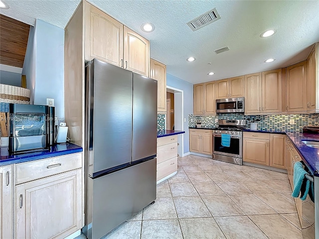 kitchen featuring stainless steel appliances, light brown cabinets, dark countertops, decorative backsplash, and a textured ceiling