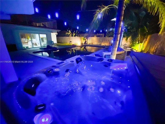 pool at night featuring a hot tub, a fenced backyard, and a patio area
