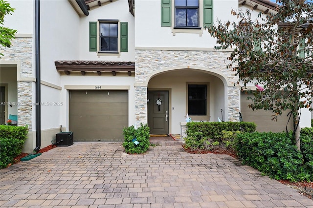 view of front of home with decorative driveway, central AC, an attached garage, stone siding, and stucco siding