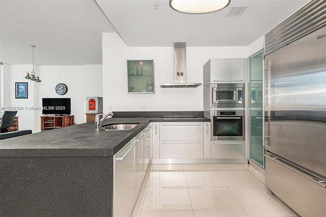kitchen featuring dark countertops, a sink, white cabinets, built in appliances, and wall chimney exhaust hood