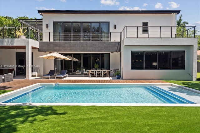 rear view of house with stucco siding, an outdoor pool, a patio area, a balcony, and a lawn