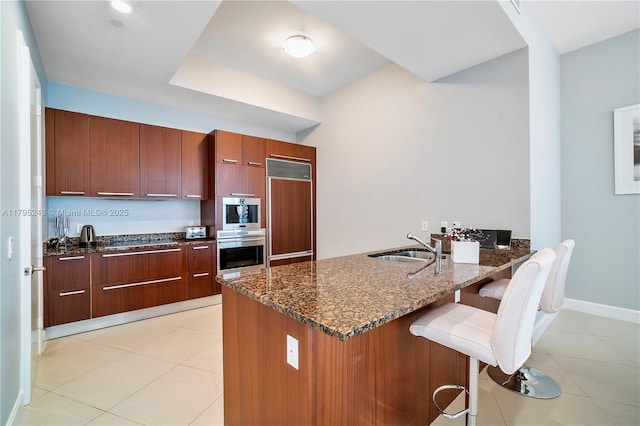 kitchen with a breakfast bar area, a sink, a peninsula, paneled built in fridge, and dark stone counters