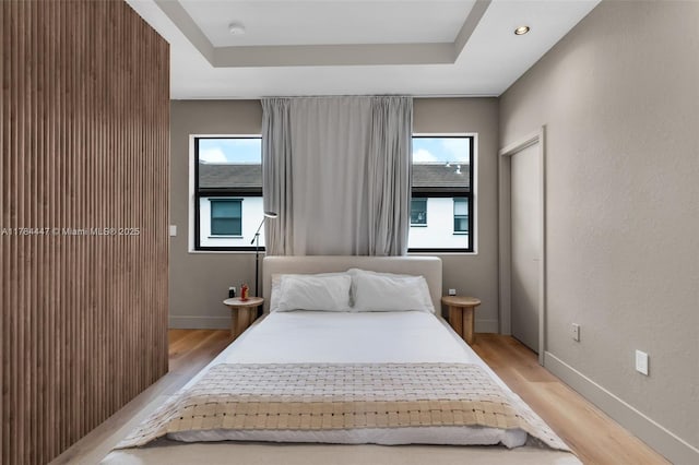 bedroom with baseboards, multiple windows, light wood-type flooring, and a raised ceiling