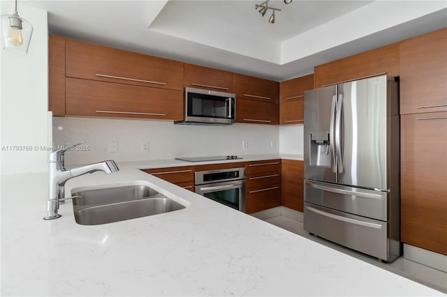 kitchen with a sink, brown cabinetry, a tray ceiling, modern cabinets, and appliances with stainless steel finishes