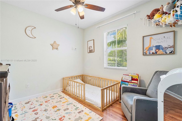 bedroom with ceiling fan, a nursery area, baseboards, and wood finished floors
