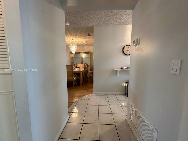hallway with an inviting chandelier, a textured ceiling, baseboards, light tile patterned floors, and visible vents