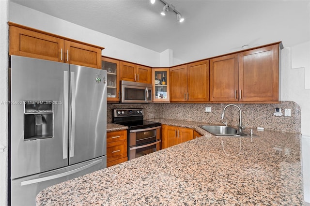 kitchen featuring brown cabinetry, light stone countertops, glass insert cabinets, a sink, and stainless steel appliances