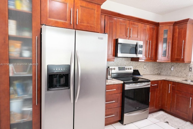 kitchen with brown cabinetry, light stone countertops, glass insert cabinets, light tile patterned floors, and stainless steel appliances