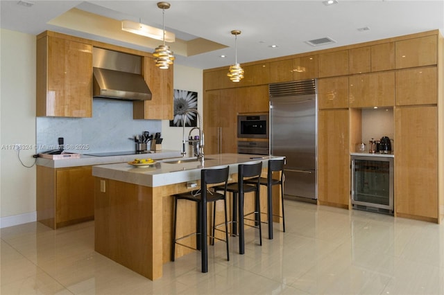 kitchen featuring wall chimney range hood, brown cabinets, beverage cooler, built in fridge, and visible vents
