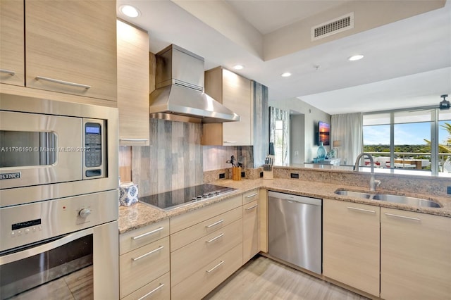kitchen with a sink, wall chimney range hood, light brown cabinetry, light stone countertops, and appliances with stainless steel finishes