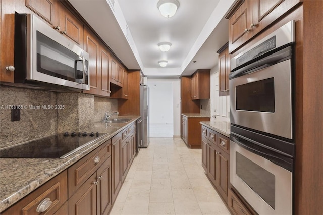 kitchen with a sink, tasteful backsplash, a tray ceiling, light stone countertops, and appliances with stainless steel finishes
