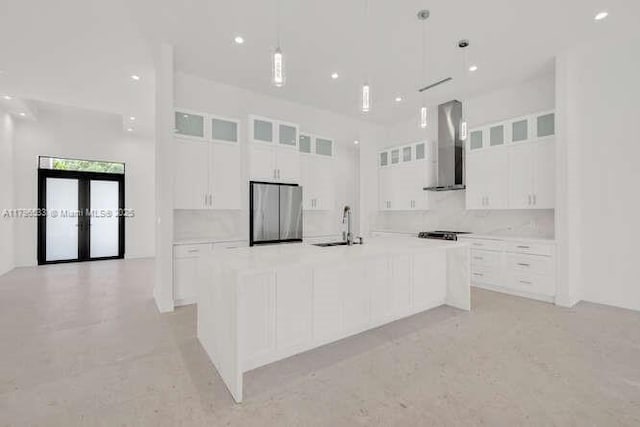 kitchen with freestanding refrigerator, wall chimney range hood, white cabinets, and a large island with sink