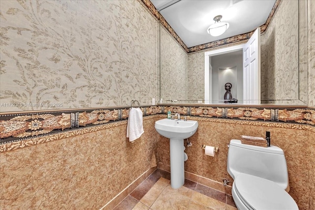 half bathroom with a sink, toilet, a wainscoted wall, tile patterned floors, and wallpapered walls