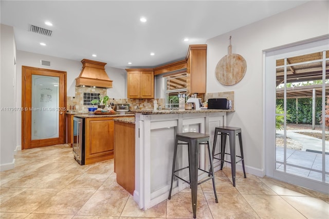 kitchen with a peninsula, visible vents, backsplash, light stone countertops, and custom range hood