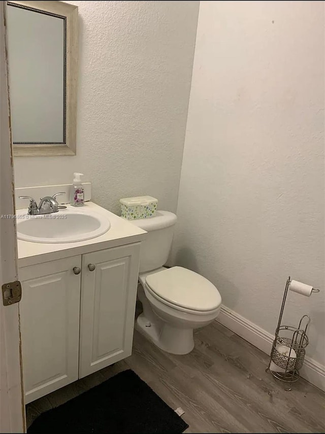 half bathroom with toilet, wood finished floors, vanity, a textured wall, and baseboards