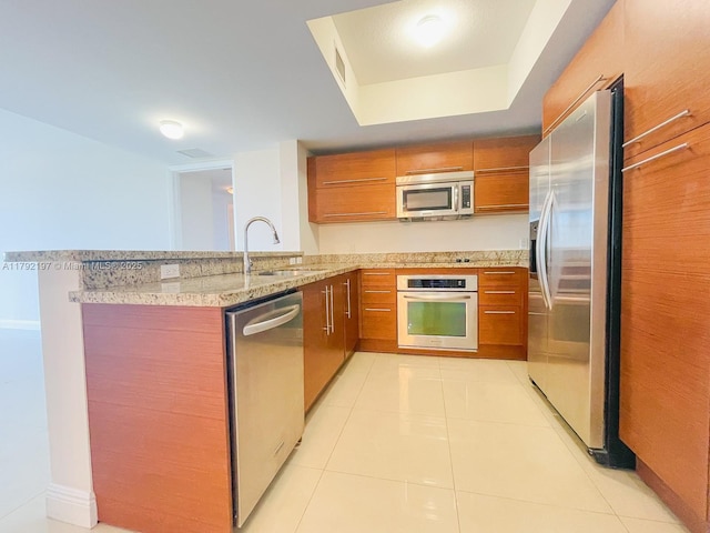 kitchen featuring a peninsula, brown cabinets, a sink, appliances with stainless steel finishes, and light stone counters