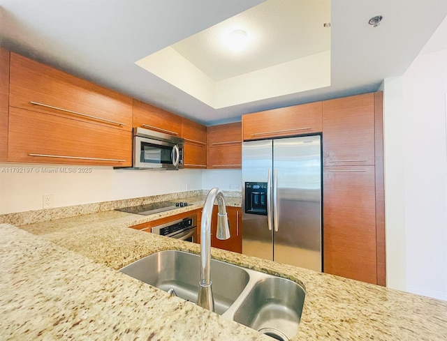 kitchen featuring brown cabinets, a sink, a tray ceiling, stainless steel appliances, and light stone countertops
