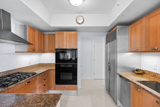 kitchen with wall chimney range hood, dark stone countertops, black appliances, light tile patterned floors, and a tray ceiling