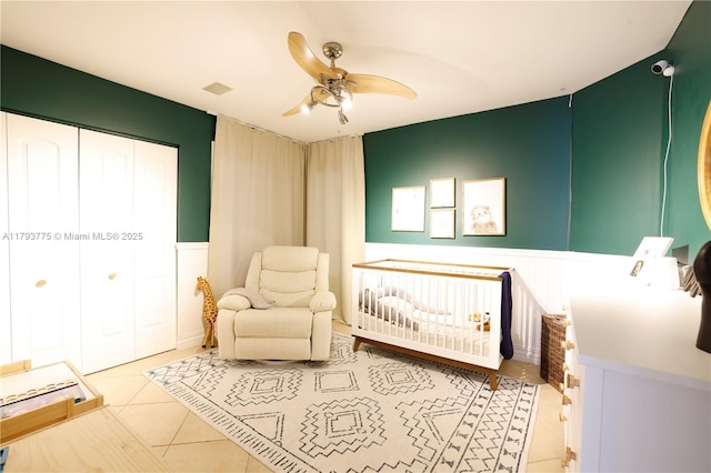 tiled bedroom featuring a nursery area, a ceiling fan, a closet, and wainscoting