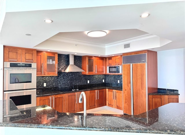 kitchen with a tray ceiling, wall chimney exhaust hood, built in appliances, and brown cabinetry