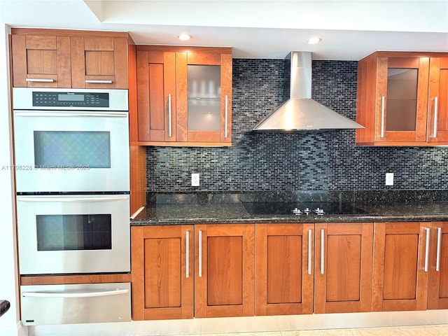 kitchen with brown cabinets, dark stone counters, multiple ovens, a warming drawer, and wall chimney range hood