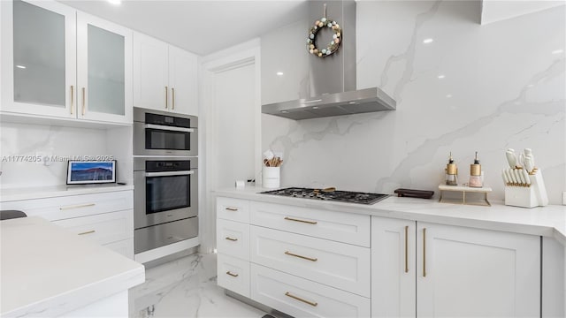 kitchen featuring wall chimney range hood, light marble finish floors, white cabinetry, stainless steel appliances, and light countertops