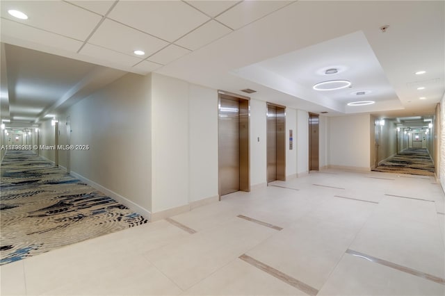 hallway with elevator, a raised ceiling, baseboards, and recessed lighting