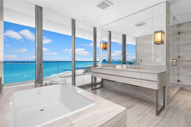 bathroom featuring view of water and beach, a tub to relax in, double vanity, a shower stall, and a wall of windows