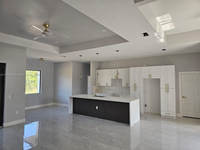 kitchen featuring a sink, a tray ceiling, baseboards, a ceiling fan, and a kitchen island with sink