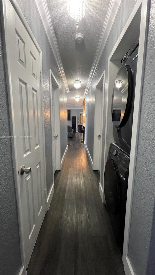 hallway with a textured wall, estacked washer and dryer, ornamental molding, a textured ceiling, and dark wood-style flooring