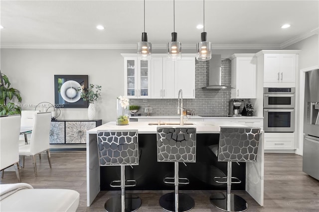kitchen featuring appliances with stainless steel finishes, wall chimney range hood, a sink, a kitchen island with sink, and light countertops