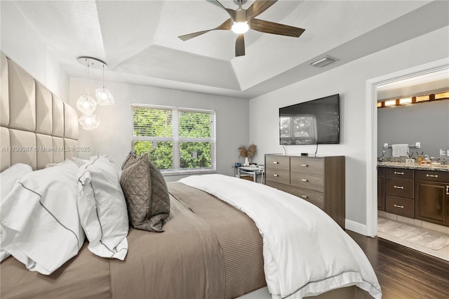 bedroom with a tray ceiling, dark wood finished floors, connected bathroom, baseboards, and ceiling fan