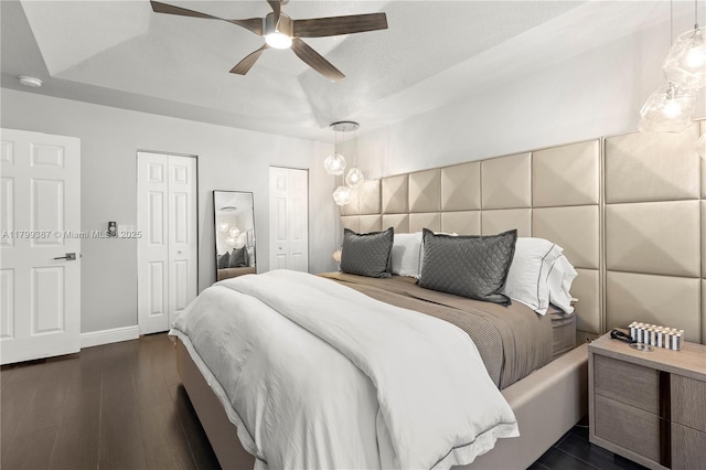 bedroom featuring a raised ceiling, multiple closets, dark wood-style flooring, baseboards, and ceiling fan