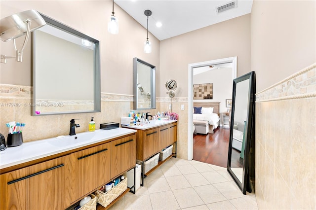 ensuite bathroom featuring tile walls, tile patterned flooring, a wainscoted wall, and two vanities