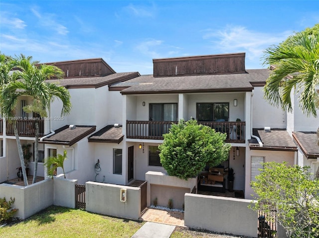 back of house with a balcony, a fenced front yard, stucco siding, and roof with shingles