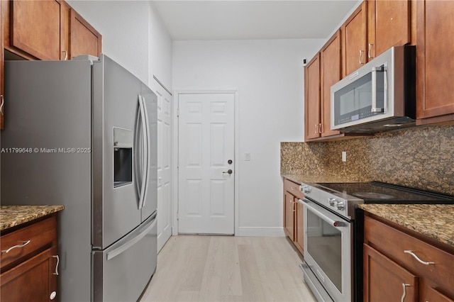 kitchen with appliances with stainless steel finishes, light wood-style floors, brown cabinets, backsplash, and dark stone countertops
