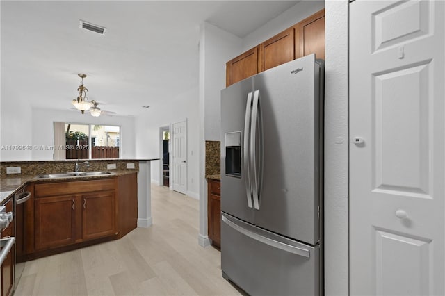 kitchen featuring stainless steel refrigerator with ice dispenser, a sink, light wood finished floors, brown cabinetry, and dark stone counters