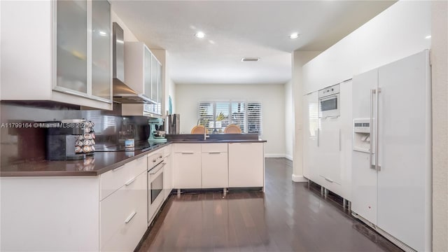 kitchen with wall chimney exhaust hood, white appliances, a peninsula, dark countertops, and recessed lighting