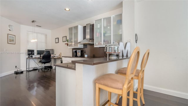 kitchen featuring wall chimney range hood, a kitchen bar, a peninsula, dark wood-style floors, and baseboards