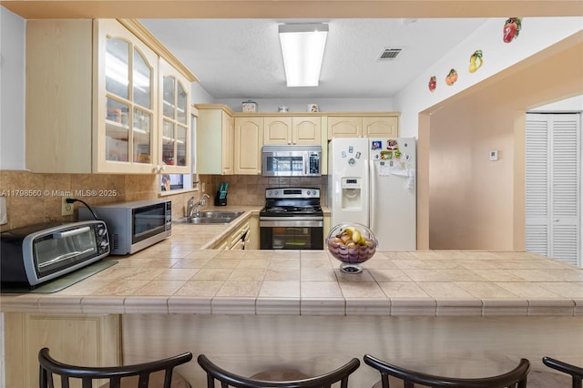 kitchen with appliances with stainless steel finishes, a sink, a peninsula, tasteful backsplash, and glass insert cabinets