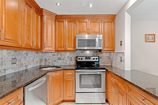 kitchen with stainless steel appliances, a sink, dark stone countertops, a textured ceiling, and brown cabinets