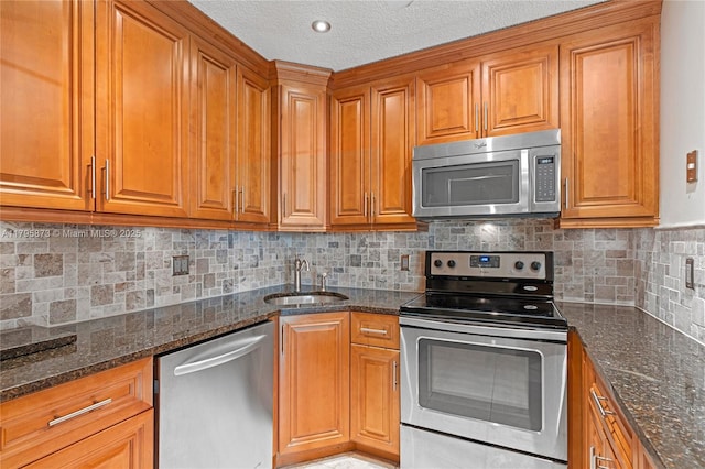 kitchen featuring stainless steel appliances, a sink, brown cabinetry, dark stone counters, and a textured ceiling