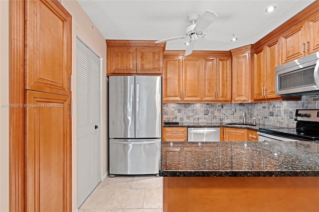 kitchen with stainless steel appliances, a sink, dark stone counters, decorative backsplash, and brown cabinetry