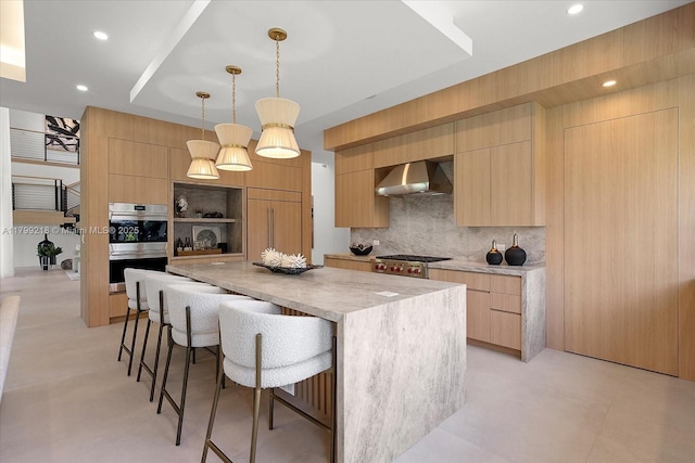 kitchen with wall chimney exhaust hood, modern cabinets, light brown cabinets, double oven, and a center island