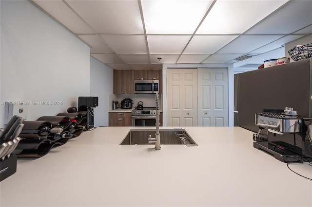 kitchen featuring stainless steel appliances, a paneled ceiling, light countertops, a sink, and brown cabinetry