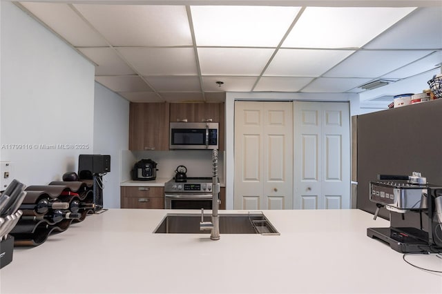 kitchen with appliances with stainless steel finishes, a paneled ceiling, a sink, light countertops, and brown cabinetry