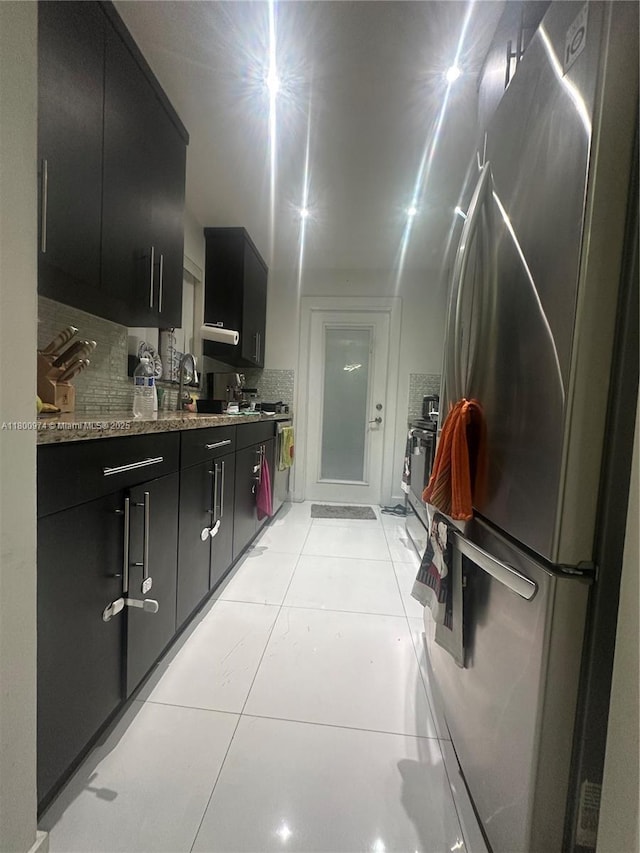 kitchen featuring freestanding refrigerator, dark cabinets, light tile patterned floors, decorative backsplash, and a sink